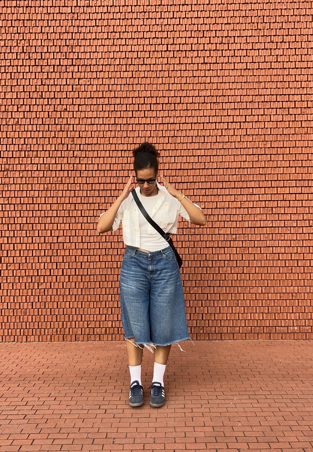Sinine denim culotte'idega, mille ääred on rebitud, valge crop top ja valge klassikaline särk. Hallid kingad ja valged sokid. Mustad prillid. Tellisseina taust.