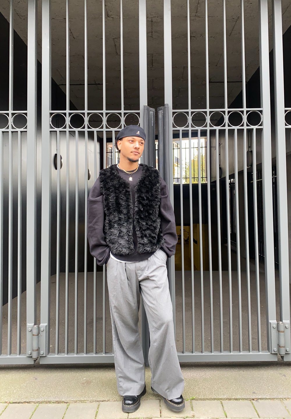 Black faux fur gilet over a black sweatshirt, grey wide-leg trousers, and black shoes, standing in front of a metal gate with vertical bars.
