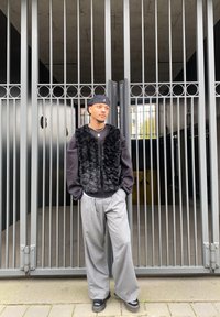 Black faux fur gilet over a black sweatshirt, grey wide-leg trousers, and black shoes, standing in front of a metal gate with vertical bars.