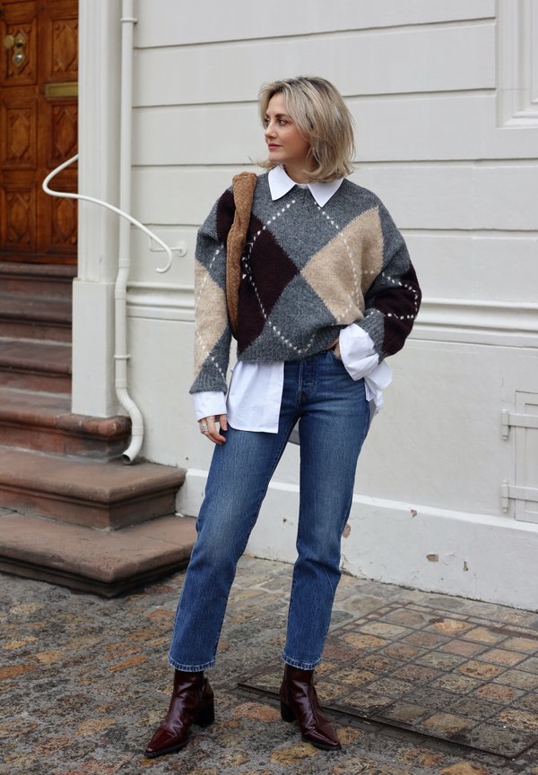 Femme debout près d'escaliers portant un pull à motif losanges par-dessus une chemise blanche, un jean bleu, des bottes marron, et tenant un sac tressé marron.