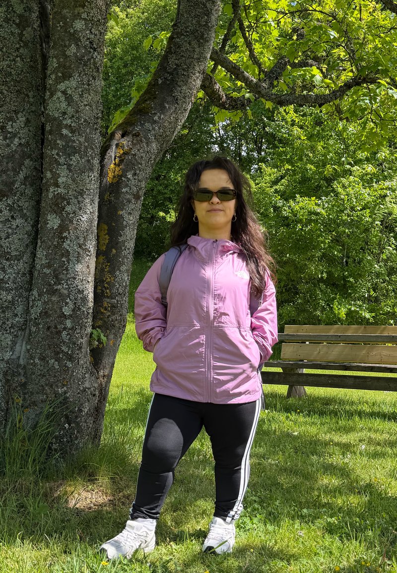 Femme portant des lunettes de soleil, une veste rose, un pantalon noir et des baskets blanches, debout les mains dans les poches près d’un arbre dans un parc vert avec un banc en bois à proximité.