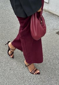 Bordeauxrote Hosen mit weitem Bein und Riemchen-Sandalen mit spitzen Absätzen. In einer Hand wird eine passende bordeauxrote Tasche gehalten. Betonhintergrund.