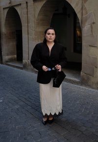 Kobieta w czarnym żakiecie i białej spódnicy trzyma okulary przeciwsłoneczne i torebkę na brukowanej ulicy, w tle kamienne łuki.