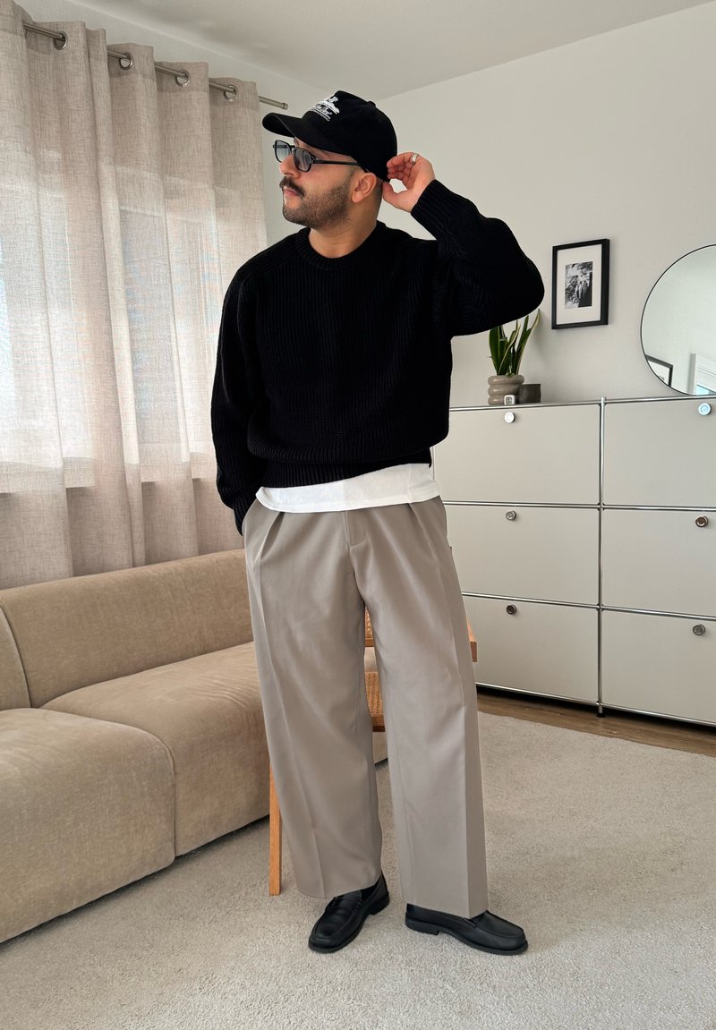 Pull en tricot noir, sous-vêtement blanc, pantalon large gris, et chaussures noires. Lumière du soleil à travers des rideaux transparents ; décoration intérieure neutre.