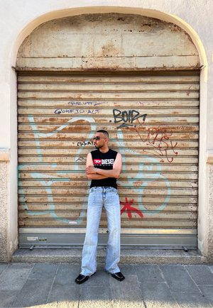 Camiseta negra sin mangas con el logo "DIESEL", jeans baggy de color azul claro y botas negras, de pie contra un fondo de metal color óxido con graffiti.