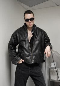 Casaco bomber de couro preto com fecho de zíper, dois bolsos frontais e gola alta. Usado sobre uma camiseta preta e combinado com jeans escuros.