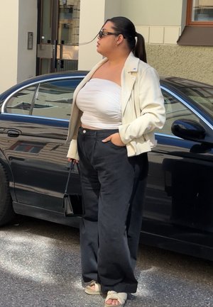 Top corto blanco, chaqueta beige clara, pantalones de pierna ancha negros y sandalias claras. Bolso negro. De pie al lado de un coche negro.