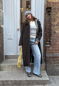 Femme debout sur des marches devant une porte, portant un long manteau sombre, un bonnet gris, des hauts superposés, un jean large, des bottes noires, tenant un sac à main jaune.