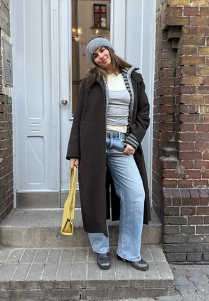 Mujer de pie en las escaleras frente a una puerta, vistiendo un abrigo largo oscuro, gorro gris, capas de prendas superiores, jeans anchos, botas negras, sosteniendo un bolso amarillo.