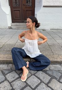 Balta be rankovių bluzonė su volanais, derinama su tamsiai mėlynomis plačiomis kelnėmis ir šviesios spalvos šlepetėmis, sėdinti ant akmenimis grįsto šaligatvio.