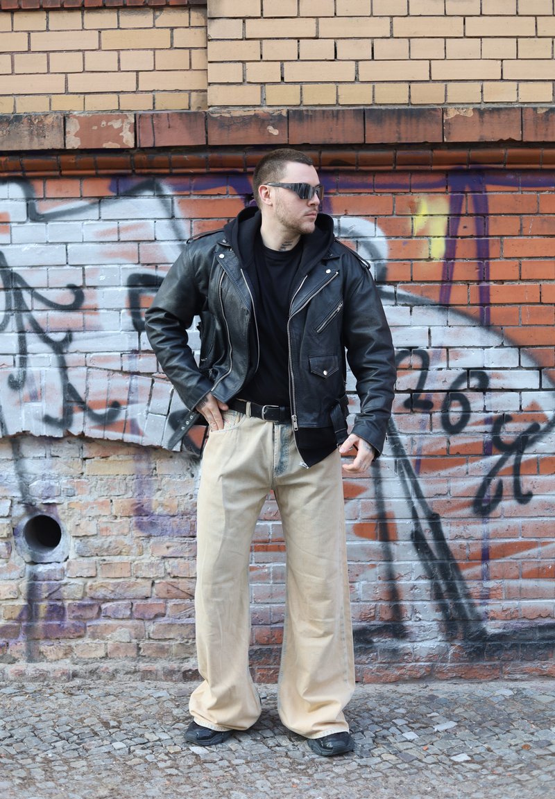 Hombre con chaqueta de cuero negra, gafas de sol y pantalones anchos beige de pierna ancha de pie contra una pared de ladrillos cubierta de grafiti.