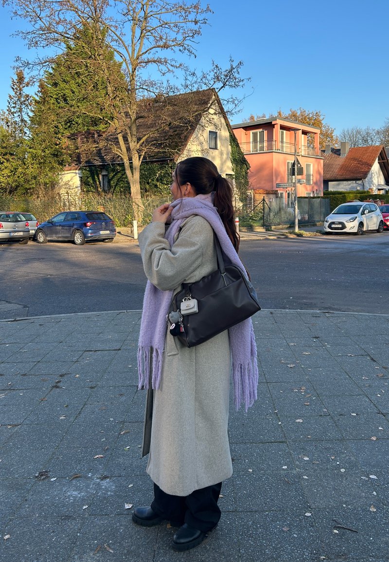 Woman in beige coat and purple scarf adjusting face mask, standing on sidewalk near parked cars and residential houses on a clear day.
