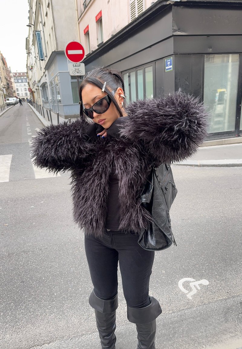 Femme portant une veste en fausse fourrure noire, des lunettes de soleil et des bottes, debout dans une rue urbaine près des panneaux d'interdiction d'entrée et de stationnement pour personnes handicapées.