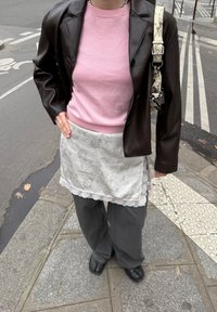 Brown leather blazer over a pink jumper, paired with a lace-trimmed white skirt and grey wide-leg trousers. Black flat shoes complete the outfit.
