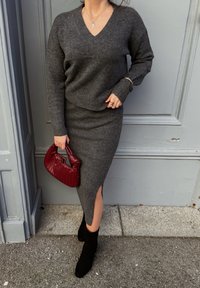 Pull en maille gris avec col en V, associé à une jupe grise assortie avec une fente latérale. Bottines noires et sac à main tissé rouge complètent le look.