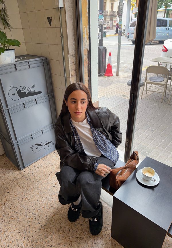 Ung kvinde iført læderjakke, der sidder ved et vindue i en café, med kaffe på et sort bord og stablede grå kasser med tegneseriedesigns i nærheden.