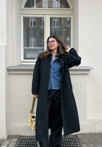 Black coat, denim shirt, black trousers, and a yellow bag with a strap. Glasses and wavy hair. Wall and window in the background.