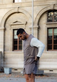 Sportos ruházatban és okosórával rendelkező férfi állít egy mellényt a szabadban, egy íves ablakokkal rendelkező történelmi épület közelében, a földön lehullott levelekkel.