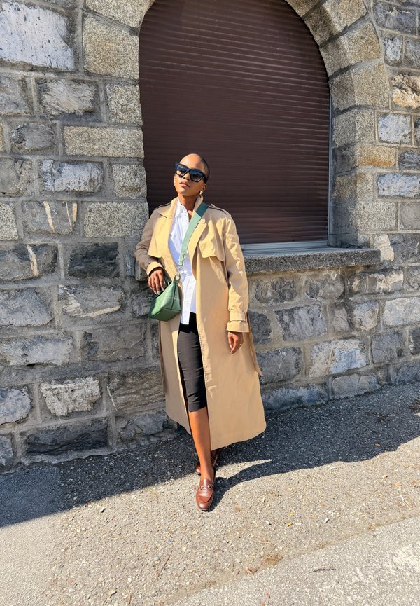 Žena v béžovom trenčkot-e, čiernych slnečných okuliaroch, bielej košeli, čiernych nohaviciach a hnedých topánkach, opierajúca sa o kamennú stenu pod zatvoreným oknom.