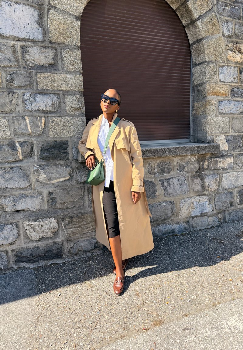 Donna che indossa un trench beige, occhiali da sole neri, camicia bianca, pantaloni neri e scarpe marroni, appoggiata a un muro di pietra sotto a una finestra chiusa.