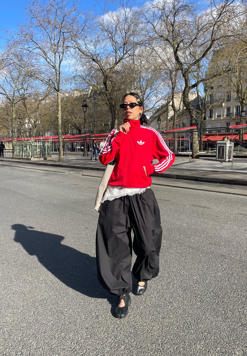 Tânăr îmbrăcat într-o jachetă roșie Adidas și pantaloni negri largi, mergând pe o stradă din oraș, cu copaci fără frunze și clădiri pe fundal.