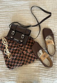 Sac bandoulière en cuir marron foncé, tissu à carreaux marron et noir avec logo, et paire de chaussures en cuir marron à lacets sur tissu beige texturé.