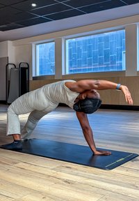 Svart yogamatta på trägolv, person i grå åtsittande outfit som utför en nedåtgående stretch, fönster i bakgrunden.