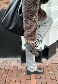 Ruske ankelstøvler med en spænde detalje, en struktureret overflade og en kraftig sål. Båret med grå jeans og en sort taske mod en murstensbaggrund.