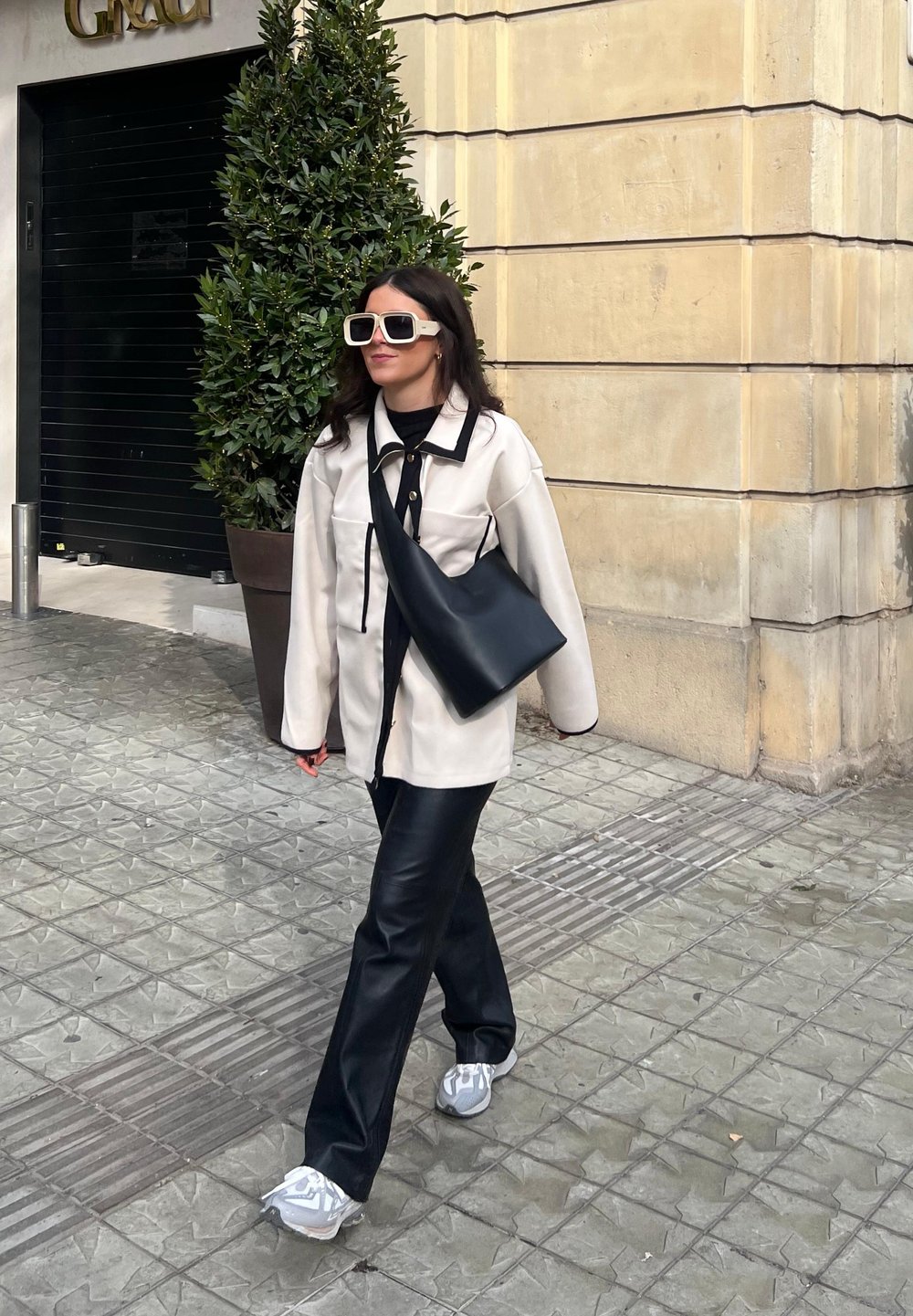 Υπερμεγέθης κρεμ ζακέτα με μαύρες λεπτομέρειες, μαύρο δερμάτινο παντελόνι, μαύρη τσάντα ώμου και λευκά γυαλιά ηλίου. Αθλητικά παπούτσια με γκρι λεπτομέρειες.