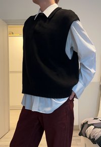 Czarny dziergany sweter bez rękawów z prążkowanym wykończeniem, nałożony na białą koszulę z kołnierzykiem, zestawiony z bordowymi spodniami. Tło utrzymane w neutralnych tonach.