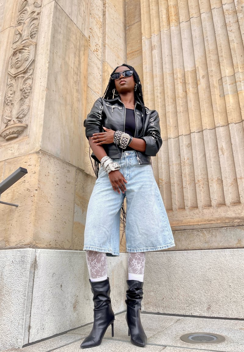 Jaqueta de couro preta, body preto, culottes de denim azul-claro, meias-arrastão e botas de salto alto pretas. De pé em frente a uma coluna texturizada.