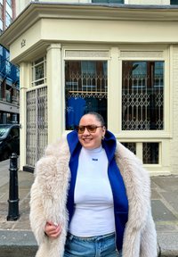 Sweat à capuche zippé bleu, col roulé blanc, manteau crème duveteux, jean en denim. Lunettes de soleil marron et boucles d'oreilles. Bâtiment à façade blanche en arrière-plan.