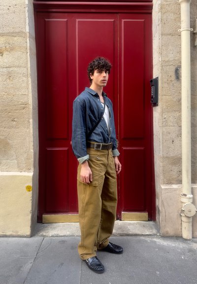 Joven con camisa azul oscuro y pantalones caqui está delante de una gran puerta roja en una calle urbana.
