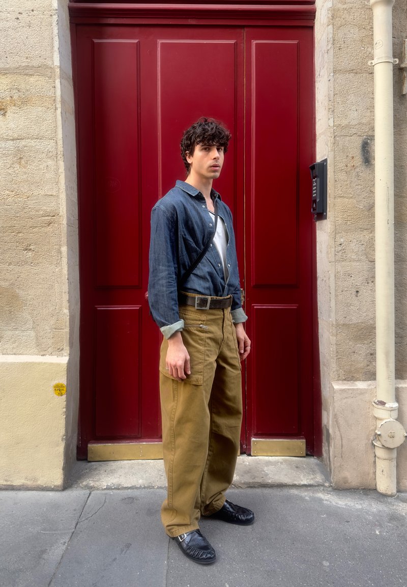 Young man wearing dark blue shirt and khaki pants stands in front of large red door on urban street.