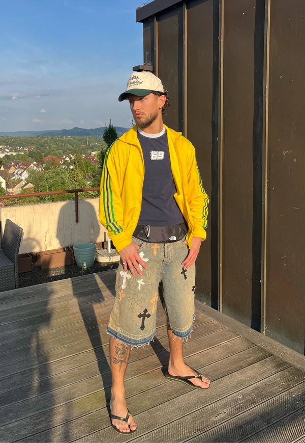 Mand i gul jakke, navy skjorte, dekorerede denimshorts, hvid kasket og flipflops står på tagterrassen med by og bakker i baggrunden.