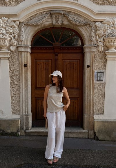 Top de tirantes de punto ligero, pantalones de lino anchos blancos y sandalias beige. El fondo presenta puertas de madera ornamentadas y una pared de piedra texturizada.