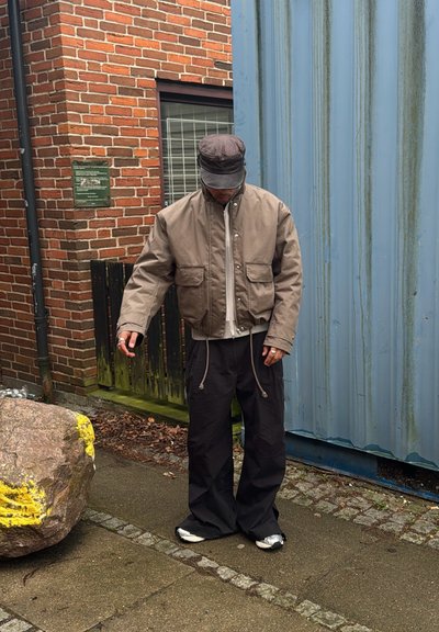 Chaqueta bomber marrón con cremallera frontal y bolsillos con solapa, combinada con pantalones negros sueltos. Fondo de piedra texturizada y paredes gris-azul.