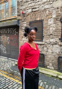 Piros kardigán, sárga felső és fekete melegítőnadrág fehér csíkokkal. Sötét haj két kontyban. Kőfal és fémredőny a háttérben.