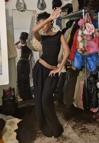 Woman in black top and wide-leg trousers posing with hand raised, standing in a room with a mirror, hanging bags, and colourful jackets.