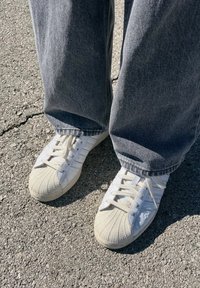 Λευκά αθλητικά παπούτσια με υφή στη μύτη, σύστημα κορδονιών και τρεις ρίγες. Φορεμένα με χαλαρούς γκρίζους τζιν παντελόνες. Φόντο από άσφαλτο.