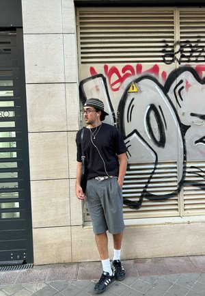 Persona que lleva una camiseta negra, pantalones cortos grises con pliegues y zapatillas negras con rayas blancas. Se incluye un sombrero a rayas y auriculares.