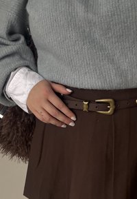 Helehall triiksärgi kraega kampsun, valge kraega särk all, pruun seelik pruunist nahkvööst kuldse pandlaga, käsi toetub vööle.