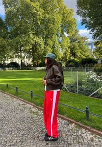 Brun læder bomberjakke, hvid skjorte, røde træningsbukser med hvide striber, grøn kasket og sorte sko, stående på en brostenssti.