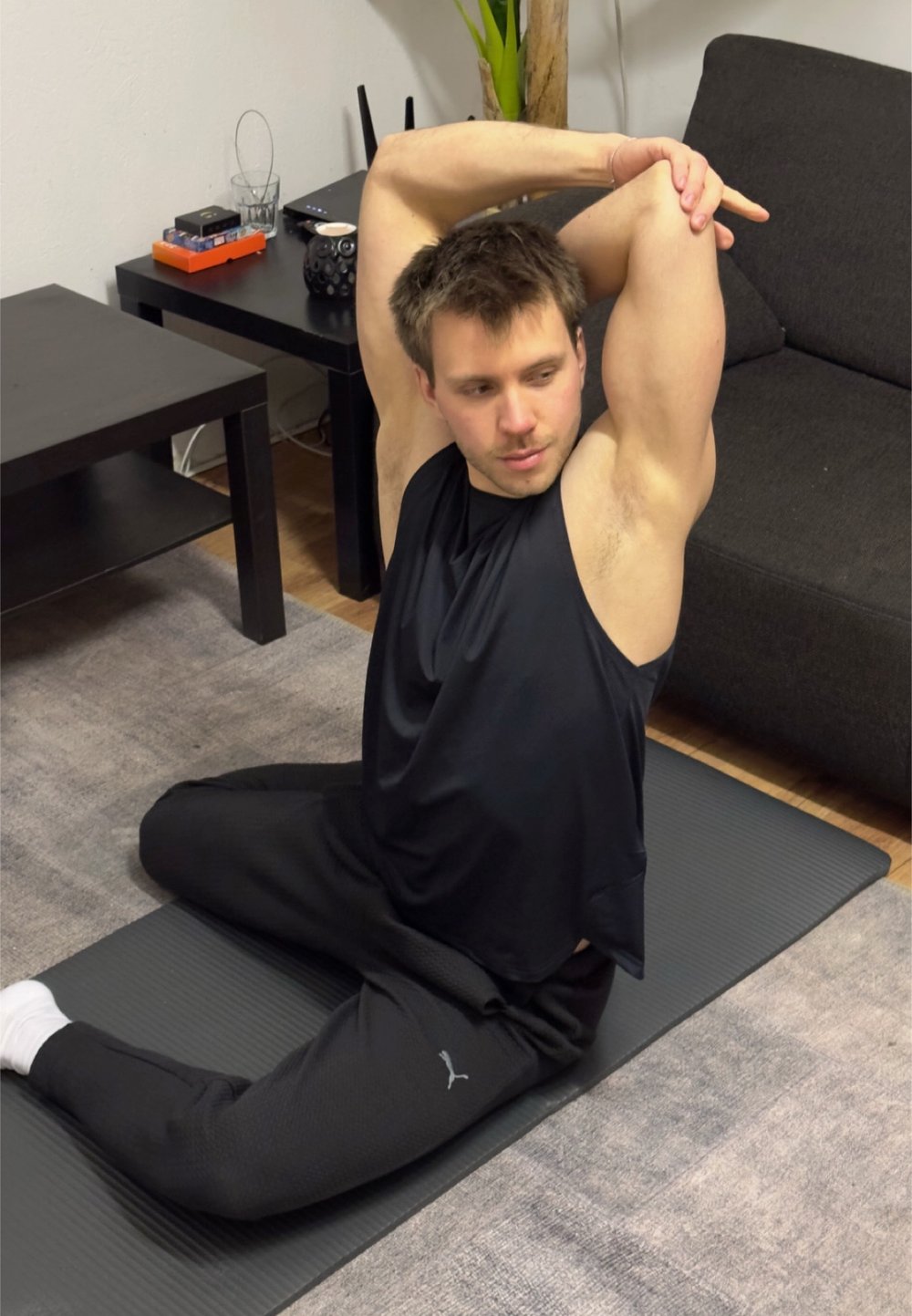 Un uomo con una maglietta senza maniche nera e pantaloni neri testurizzati esegue un allungamento seduto su un tappetino yoga nero testurizzato in un soggiorno.