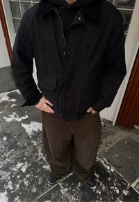 Veste noire à fermeture éclair avec un col en velours côtelé, dotée de deux poches avant et d'une coupe ajustée. Assortie à un pantalon en velours côtelé marron et des bottes noires.