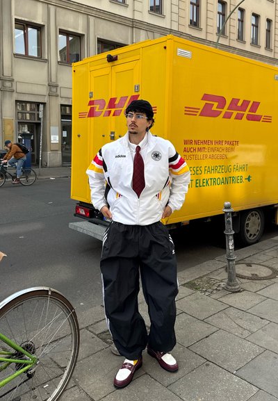Chaqueta blanca con rayas negras, rojas y amarillas, pantalones de chándal negros, zapatos marrón oscuro y una corbata. El fondo presenta un camión de entrega amarillo de DHL.