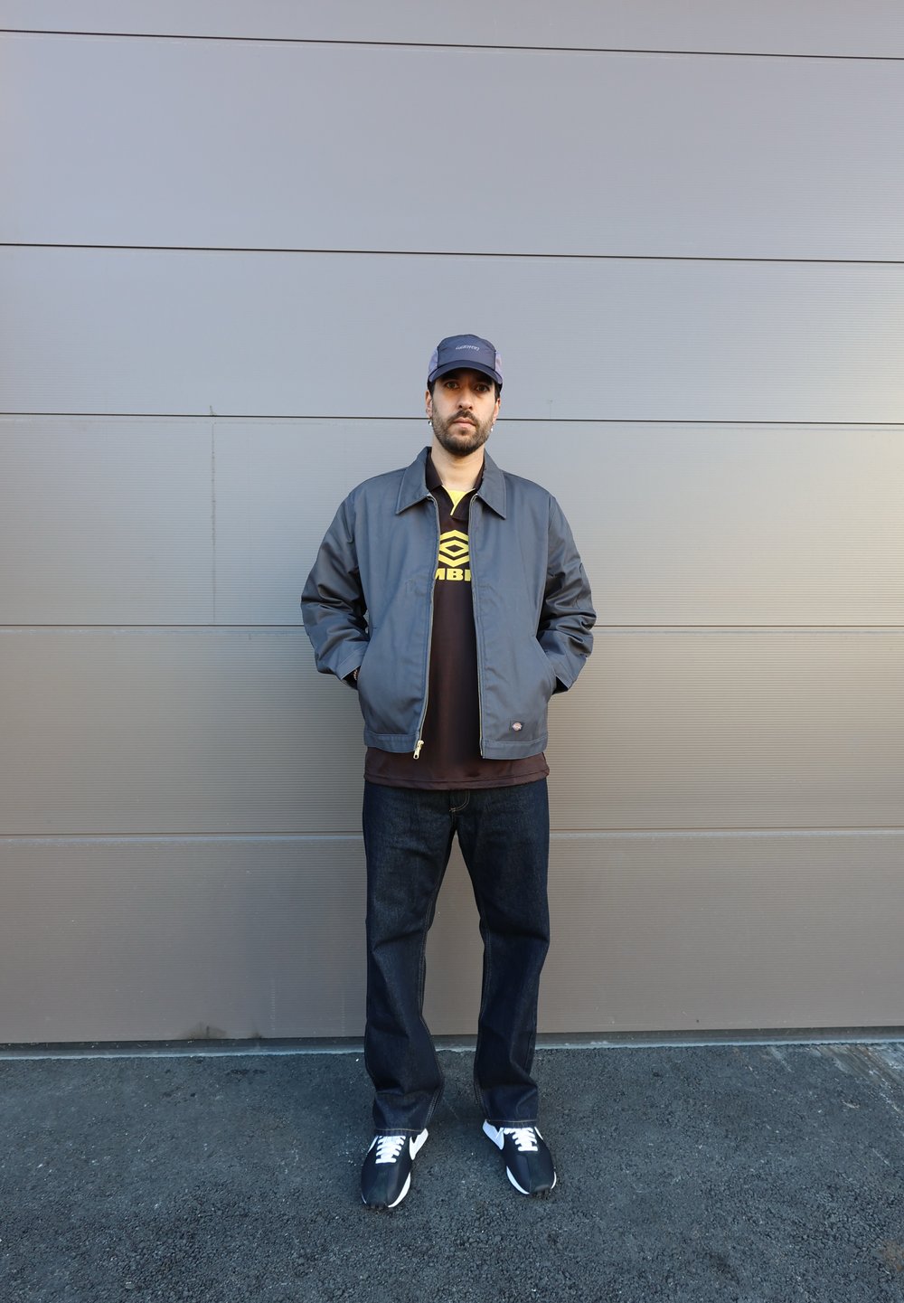 Chaqueta gris con cremallera frontal, camiseta negra con un logo, jeans azul oscuro y zapatillas negras. Diseño del uniforme, corte informal y colores sólidos.