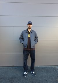 Chaqueta gris con cremallera frontal, camiseta negra con un logo, jeans azul oscuro y zapatillas negras. Diseño del uniforme, corte informal y colores sólidos.
