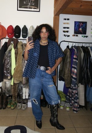 Camisa de manga corta azul con un patrón sobre una camiseta negra, jeans azules rasgados y botas vaqueras negras con bordados blancos. Los accesorios incluyen un collar de cuentas.