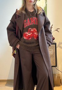 Jeune personne portant un long manteau marron, un pull foncé avec des cerises rouges et le texte « GANT », un pantalon marron à fines rayures, debout à l'intérieur près des meubles.